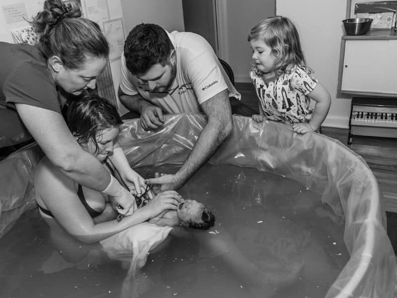 Midwife Neva Gerke helping new father cut the umblical cord at a home water birth with older sister looking on.